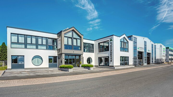 Ein modernes weißes Gebäude mit einem grauen Dach, großen Fenstern und einem Schild für "Mahnenholz" steht an einer Straßenecke, mit einem klaren blauen Himmel und Bäumen im Hintergrund.