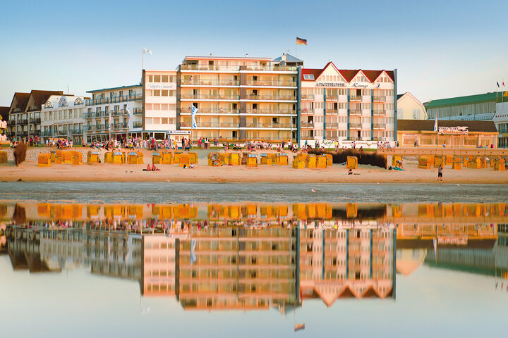 Ein Hotel am Strand, umgeben von orangefarbenen Liegestühlen und Sonnenschirmen, spiegelt sich im ruhigen Wasser, über dem eine deutsche Flagge weht.