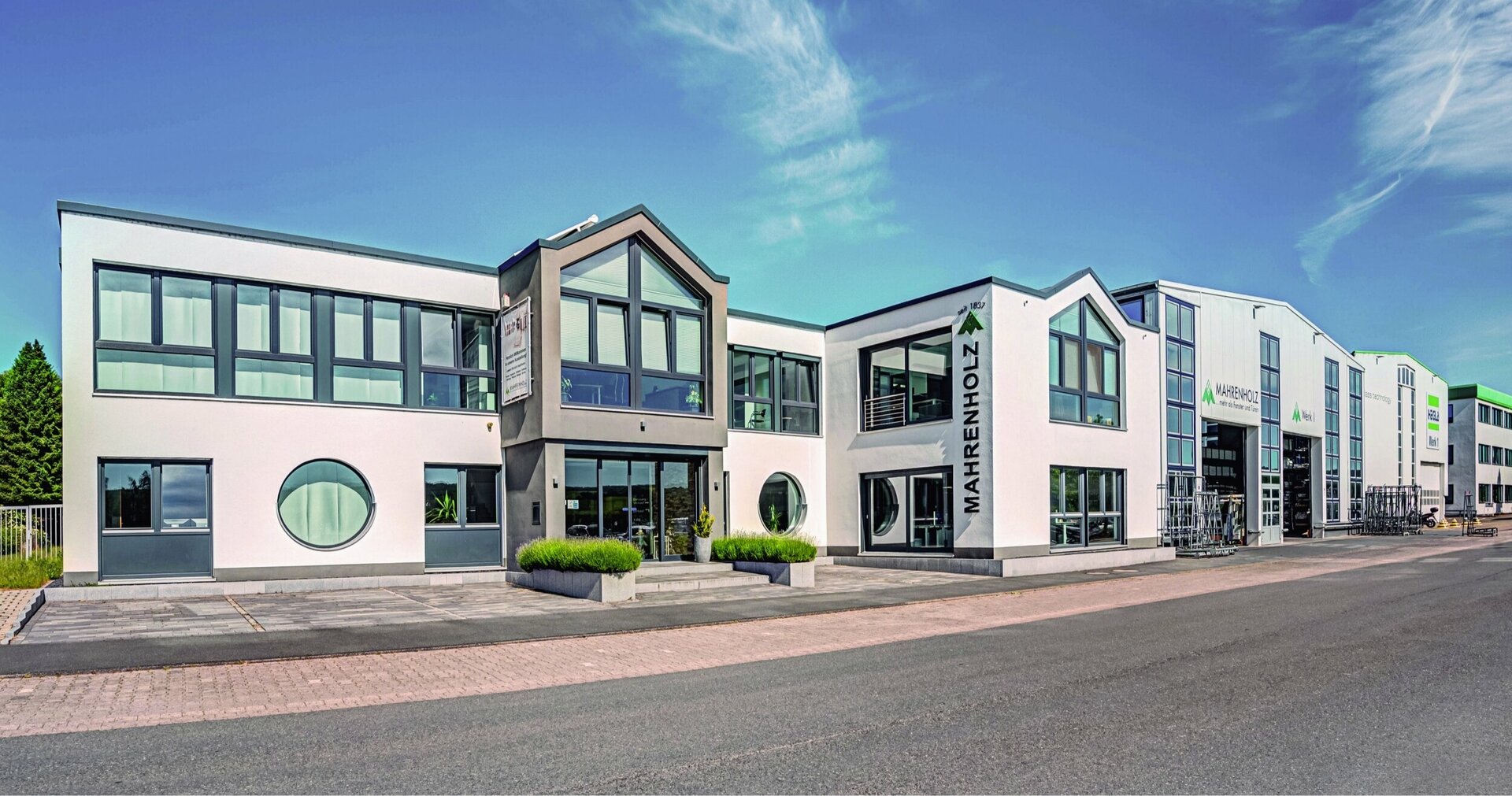 Ein modernes weißes Firmengebäude mit grauem Dach, großen Fenstern und einem Schild "Mahrenholz" steht an einer Straße, daran schließen eine Produktionshalle sowie weitere Gebäude an. Der Himmel ist blau mit einigen wenigen Wolken.
