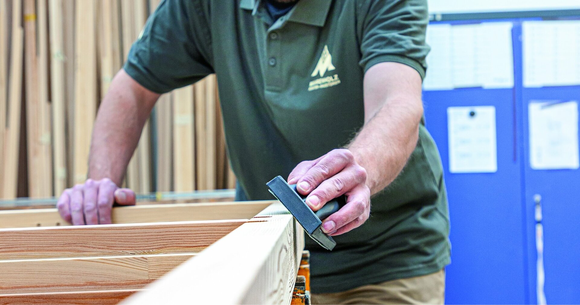Eine Person in einem grünen Polohemd mit einem Logo auf der Brust schleift mit einem schwarzen Werkzeug ein Holzbrett in einer Werkstatt mit blauen Wänden und einem Stapel von Holzbrettern.
