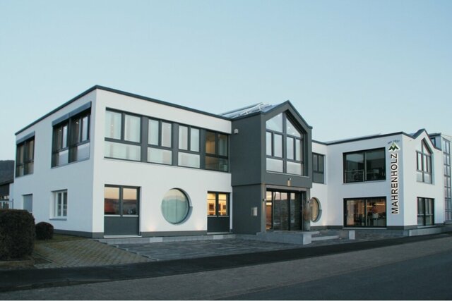 Ein modernes weißes Gebäude mit schwarzen Akzenten, großen Fenstern und einem Flachdach ist das Verwaltungsgebäude von MAHRENHOLZ  auf dessen Standort ein Schild hinweist.