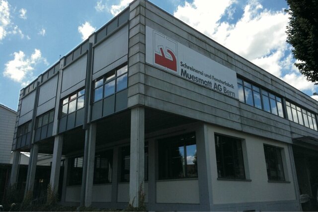 Ein modernes, graues Betongebäude mit grossen Fenstern und dem roten Logo "Schreinerei und Fensterbau Muesmatt AG Bern" steht vor einem strahlend blauen Himmel.